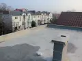 Nowy, betonowy dach płaski z kominem i widokiem na szereg domów z dachówką.