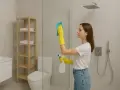 Kobieta w żółtych rękawiczkach czyści kabinę prysznicową, używając gąbki i sprayu. Dowiedz się, czym myć kabinę prysznicową, by lśniła czystością.