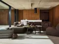 Nowoczesny salon w stylu bauhaus. Duża, narożna sofa i fotel w odcieniach brązu, betonowa podłoga, drewniane panele i szklane drzwi.