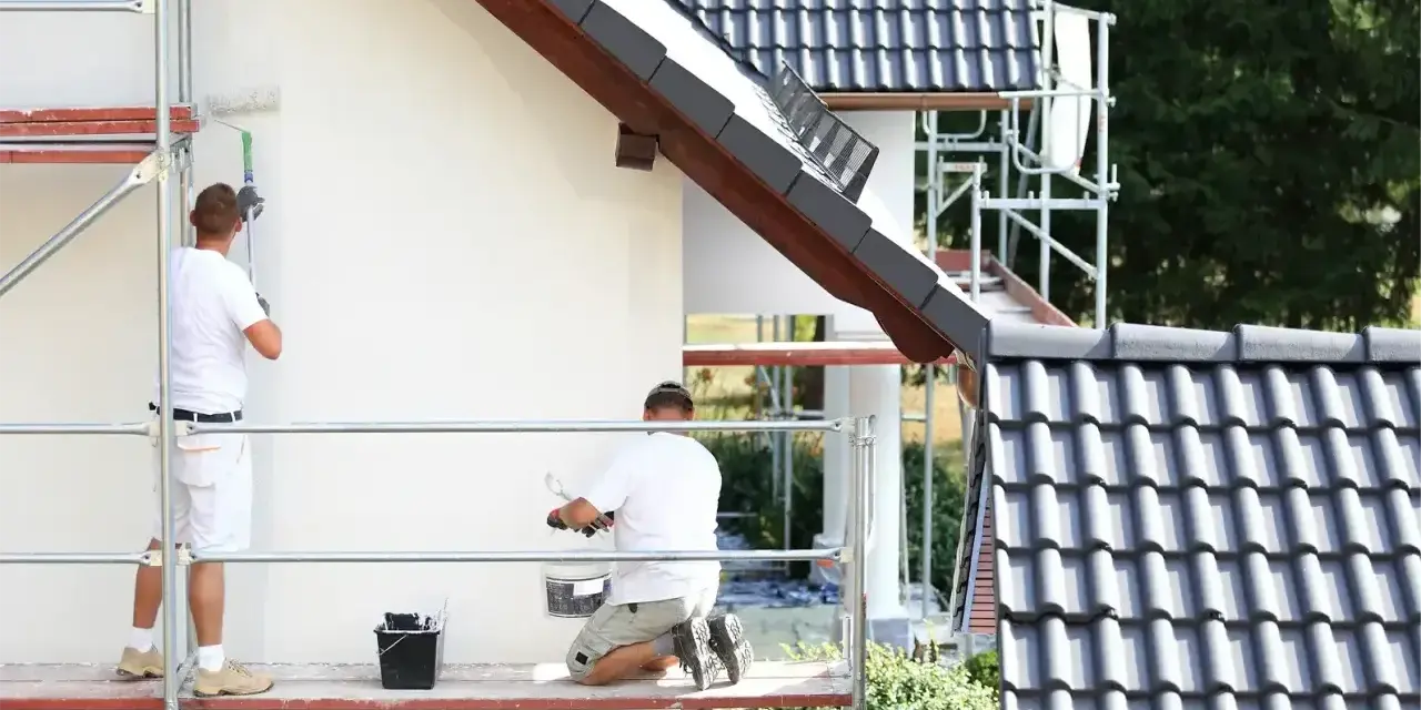 Dwóch mężczyzn na rusztowaniu wykonuje malowanie elewacji białej ściany domu.