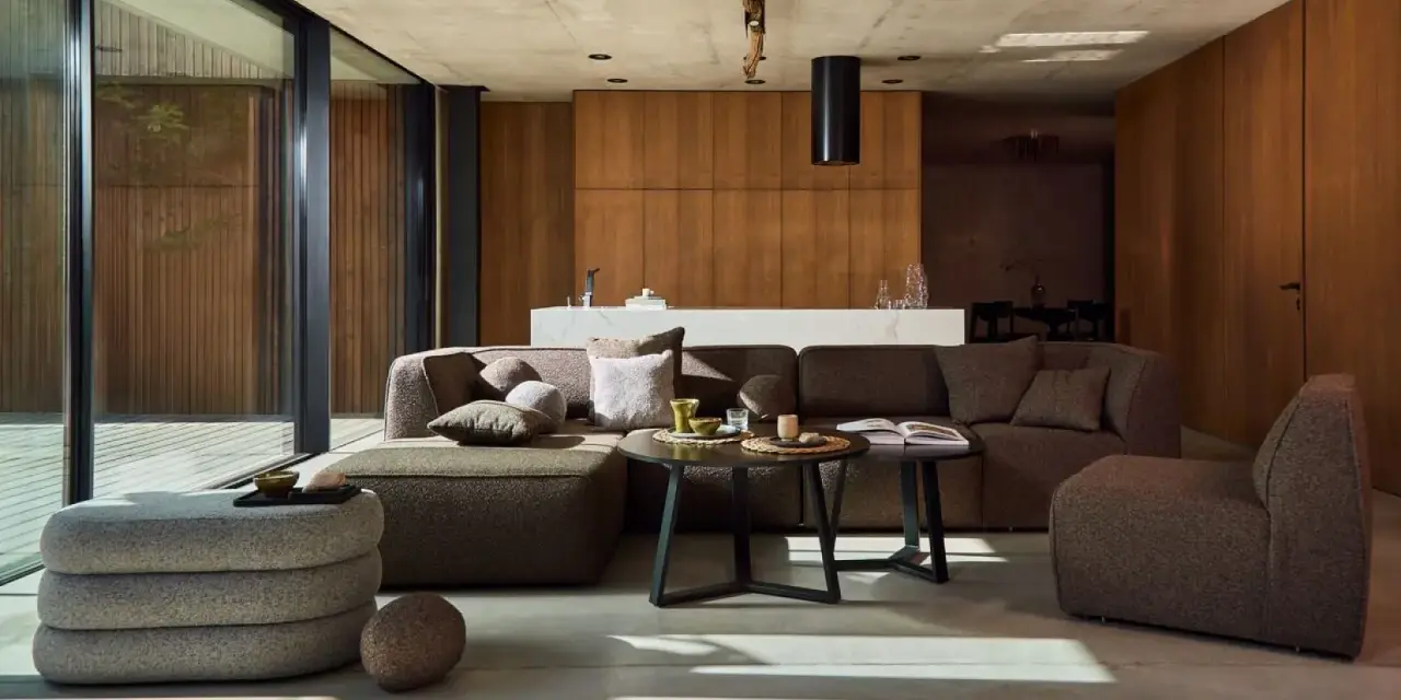 Nowoczesny salon w stylu bauhaus. Duża, narożna sofa i fotel w odcieniach brązu, betonowa podłoga, drewniane panele i szklane drzwi.
