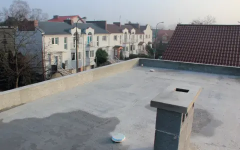 Nowy, betonowy dach płaski z kominem i widokiem na szereg domów z dachówką.