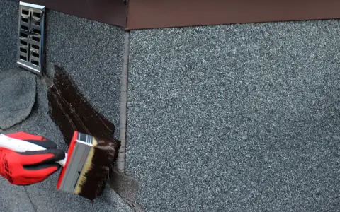 Rękawica w czerwonej rękawicy nakłada ciemną masę uszczelniającą na dach. To abizol p, który chroni przed wilgociącią.
