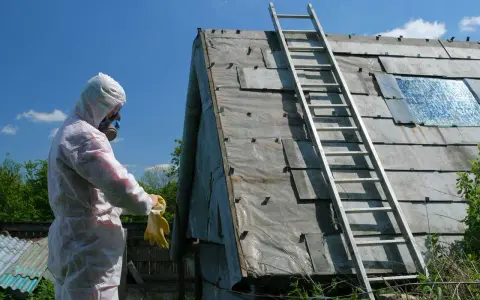 Osoba w kombinezonie ochronnym przygotowuje się do wymiany eternitu na dachu. Drabina oparta o połać dachową.
