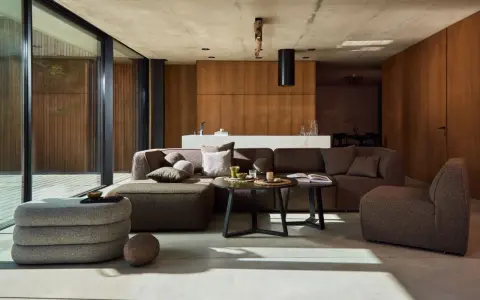 Nowoczesny salon w stylu bauhaus. Duża, narożna sofa i fotel w odcieniach brązu, betonowa podłoga, drewniane panele i szklane drzwi.
