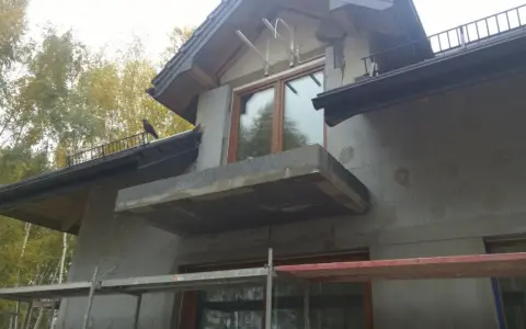 Budowa domu w jesiennej scenerii. Widać rusztowania, okna i balkon, a także ptaka na dachu. Trwają prace nad izolacją, która wpłynie na współczynnik przenikania ciepła.