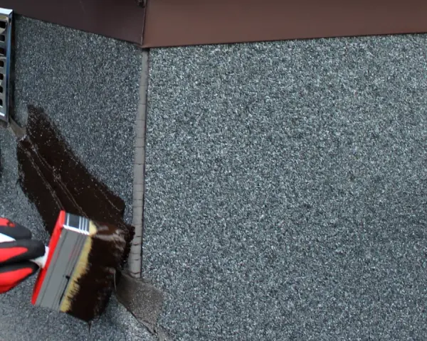 Rękawica w czerwonej rękawicy nakłada ciemną masę uszczelniającą na dach. To abizol p, który chroni przed wilgociącią.