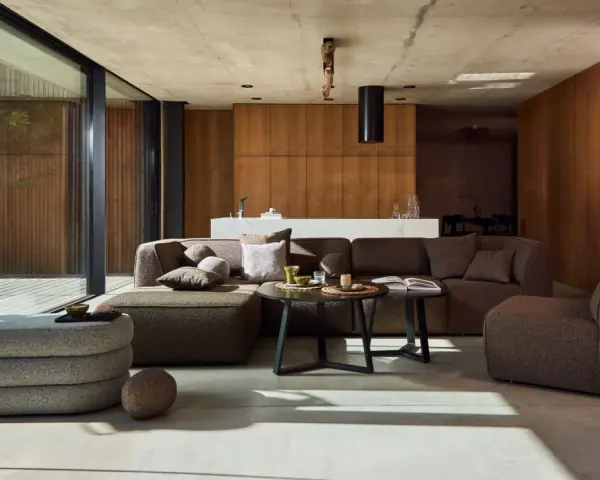 Nowoczesny salon w stylu bauhaus. Duża, narożna sofa i fotel w odcieniach brązu, betonowa podłoga, drewniane panele i szklane drzwi.