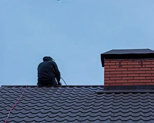 Przegląd kominiarski na dachu. Kominiarz w czarnej kurtce siedzi na ciemnym dachu, obok ceglanego komina.