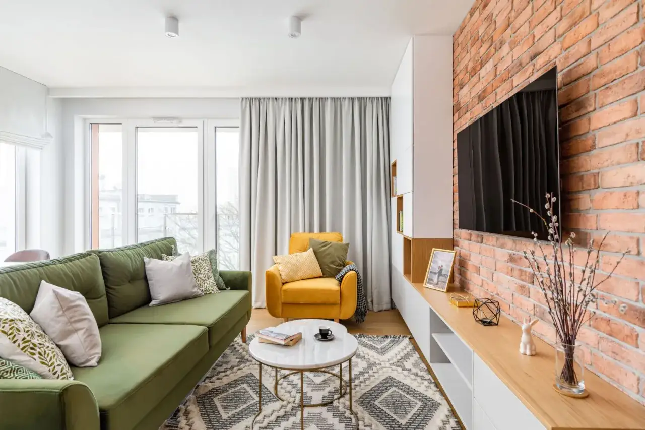 Zielona sofa i żółty fotel tworzą przytulny kącik. Inspiracja, jak urządzić salon z ceglaną ścianą i nowoczesnymi meblami.