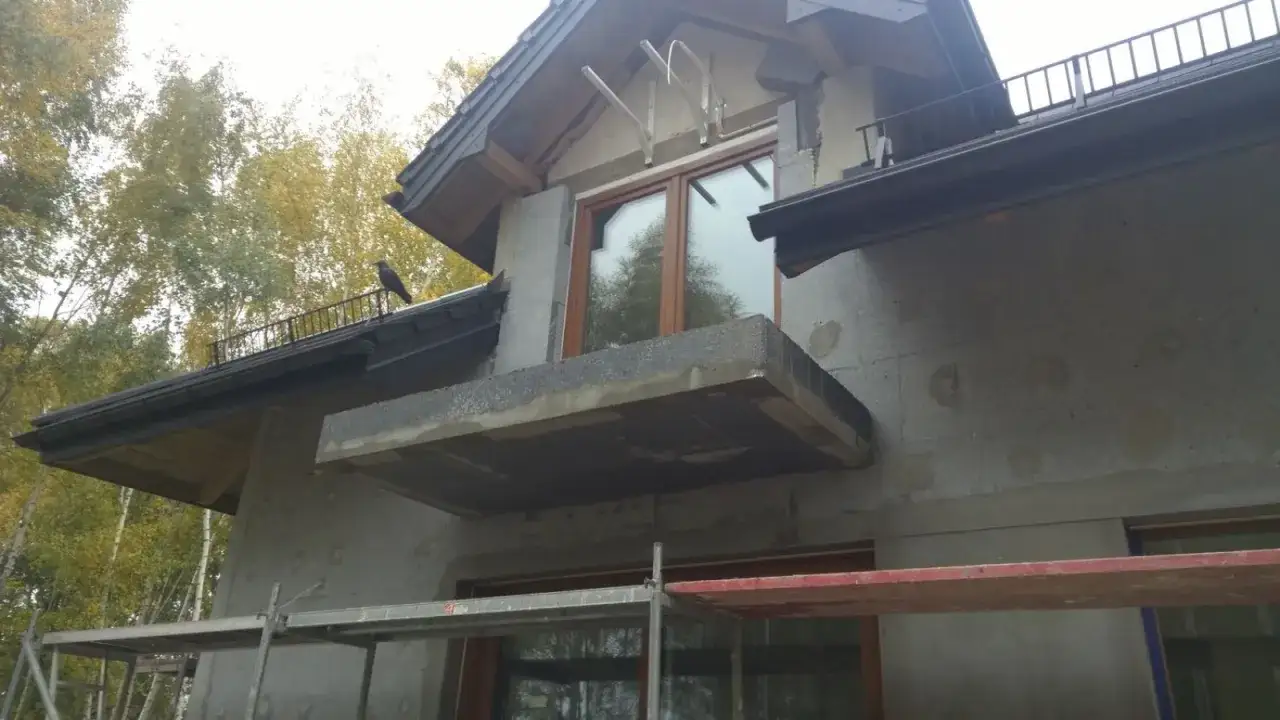 Budowa domu w toku, szara elewacja z balkonem i oknem. Na dachu siedzi ptak.