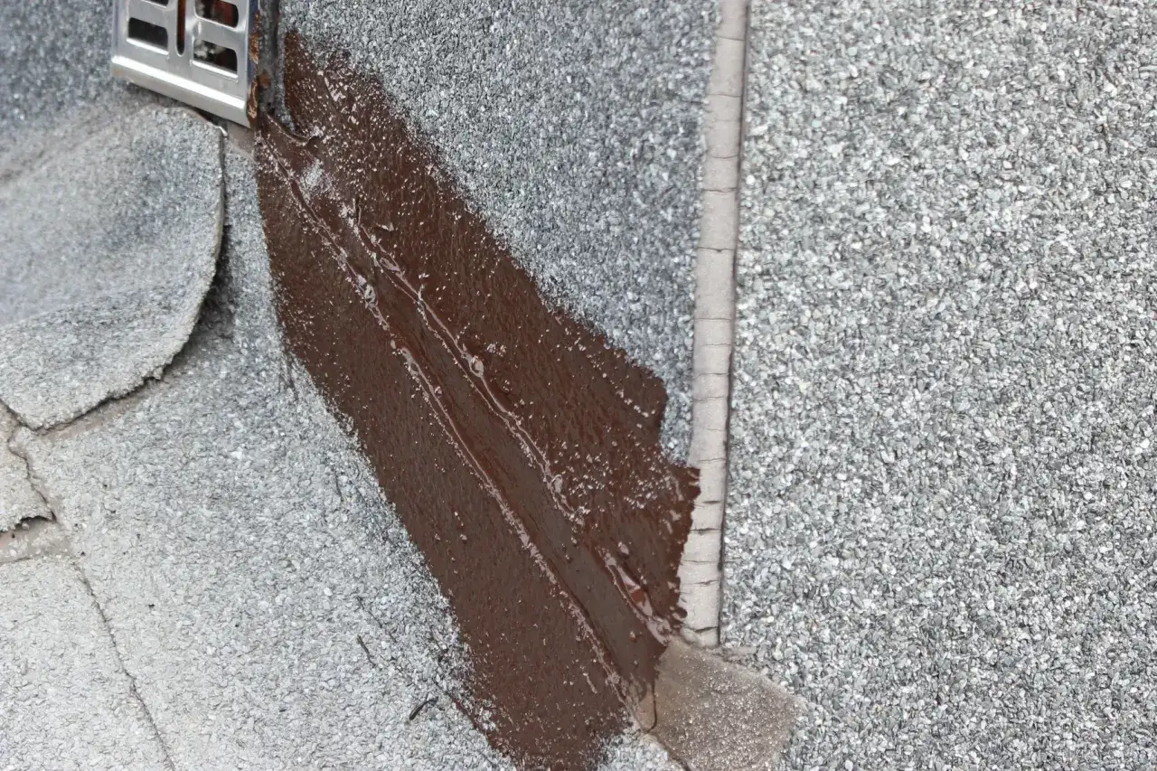 Fragment dachu pokryty papą, z widoczną brązową masą bitumiczną uszczelniającą połączenie i metalowym elementem odpływowym.