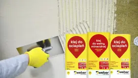 Rękawica w żółtej rękawicy nakłada klej na ścianę. Obok worki z napisem "klej do ociepleń" – idealny, by przykleić styropian do blachy.